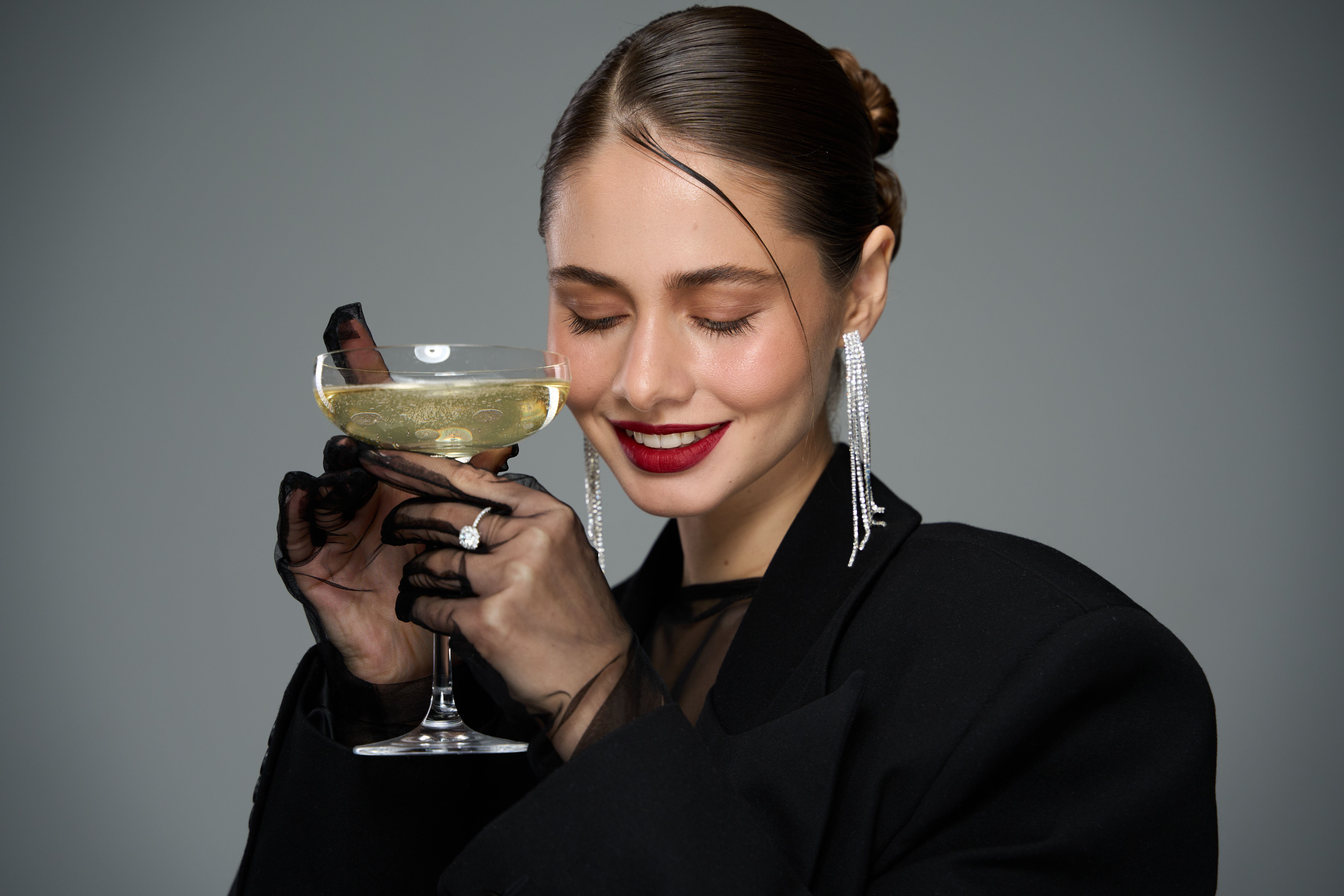 Portrait of a woman with sleek hair and bold red lipstick, wearing a black blazer and holding a cocktail glass, smiling softly against a clean studio background.
