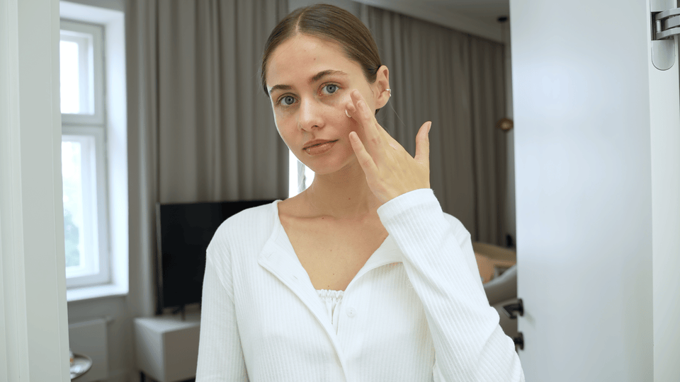 Woman applying skincare to her cheek with her fingertip in a bright indoor setting, demonstrating a simple facial routine.