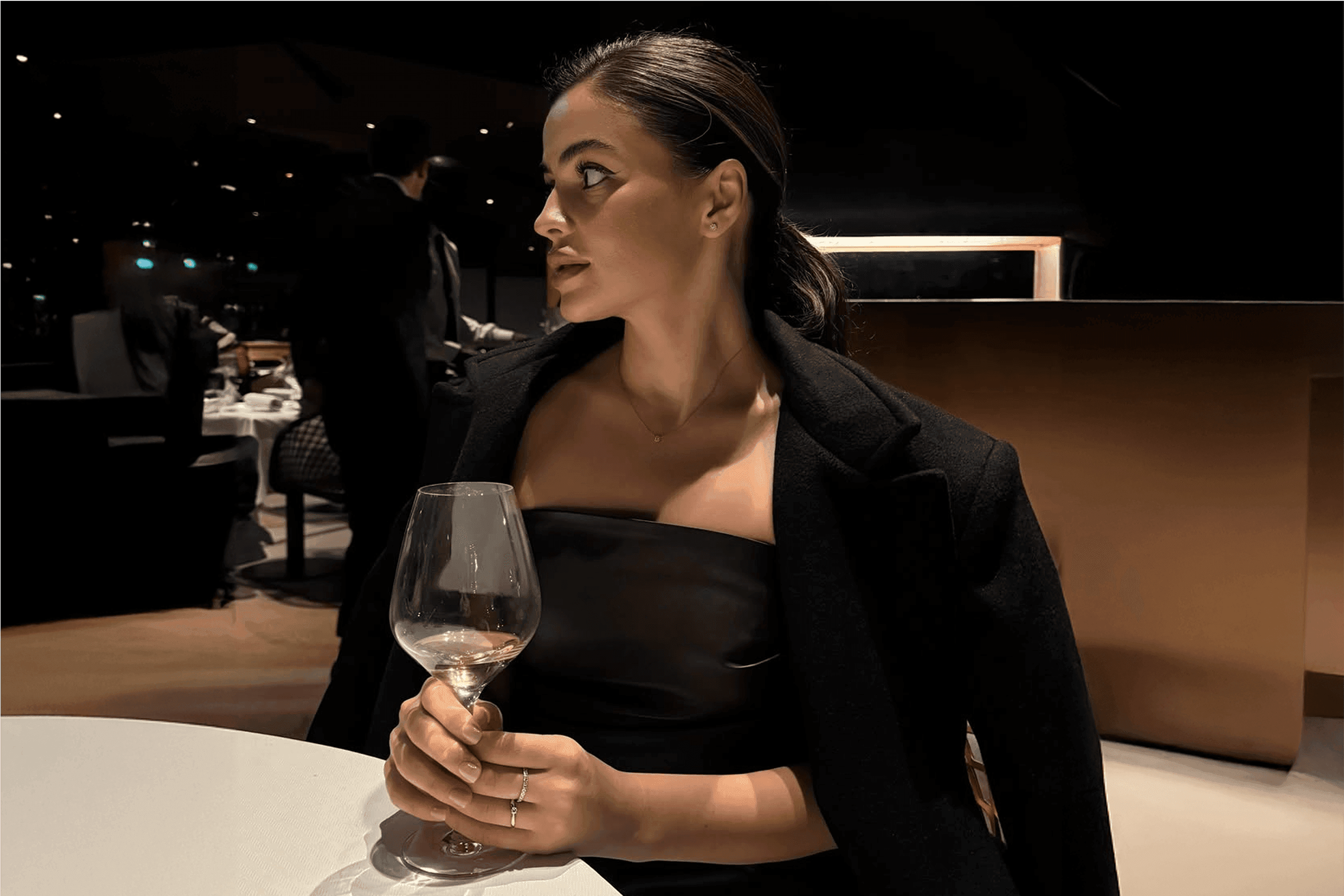 Woman in an elegant evening setting holding a wine glass, wearing a black outfit with sleek hair and refined makeup, looking to the side in a softly lit restaurant interior.