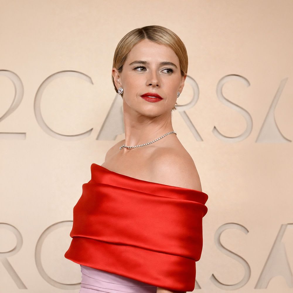 Jessie Buckley on the Oscars red carpet wearing a bold red off-shoulder satin gown, styled with sleek side-parted hair, classic red lipstick, soft eye makeup, and delicate diamond jewelry against a warm beige backdrop.