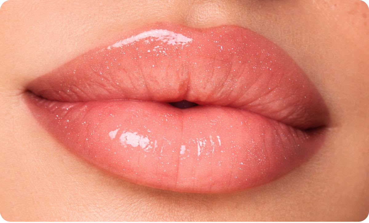 Close-up of full lips with a glossy, shimmering finish, featuring a soft peach-pink tone and light-reflecting shine concentrated at the center for a plumping effect.