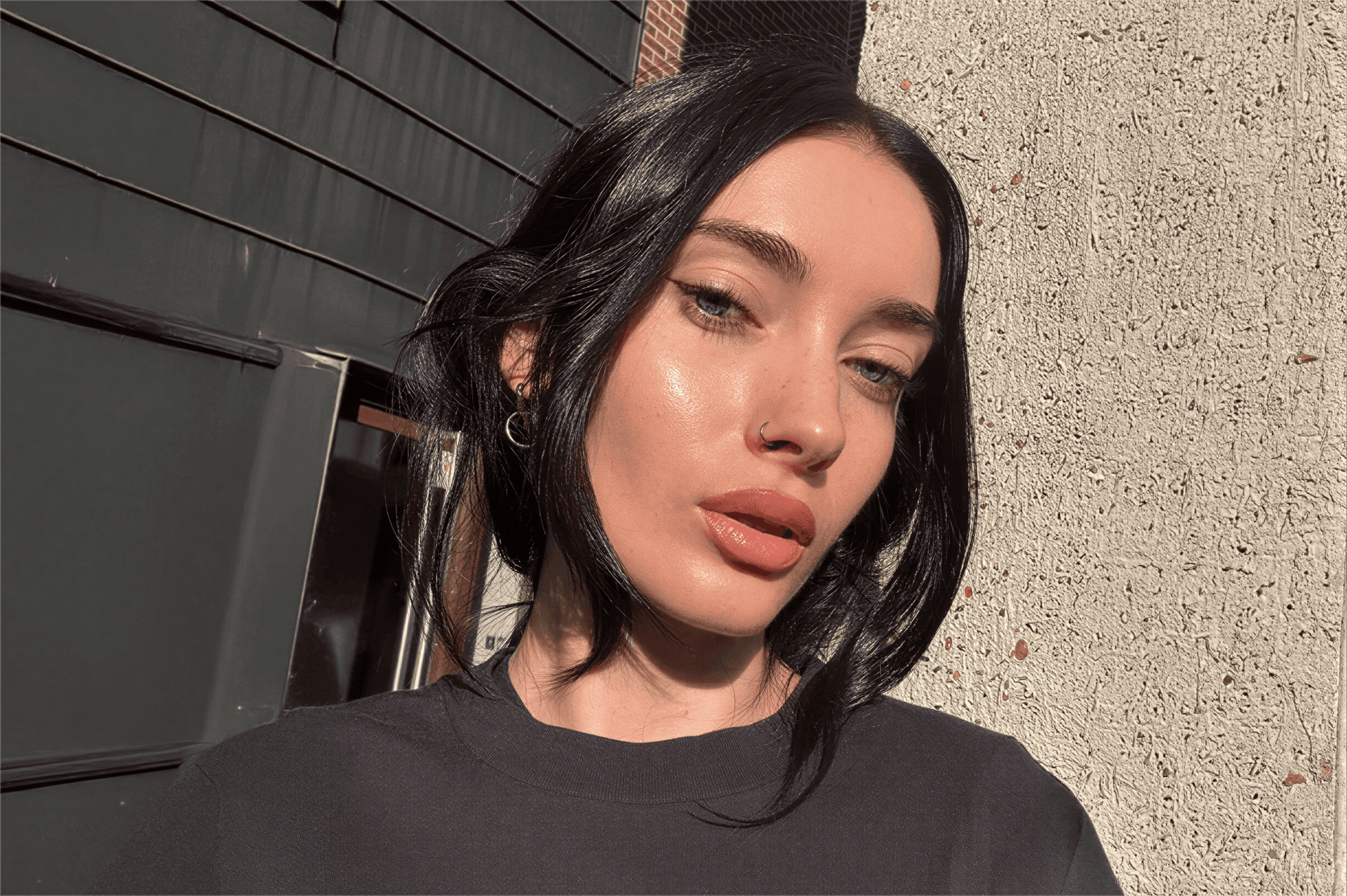 Close-up of a woman with short dark hair in sunlight, wearing minimal makeup with softly defined eyes and nude lips, highlighting natural skin texture and glow.