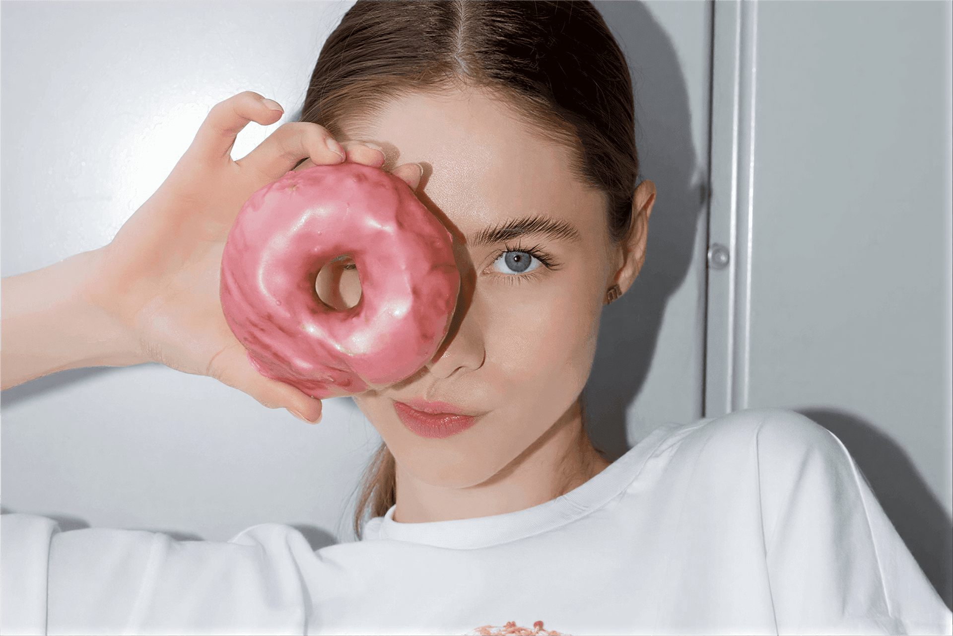 Woman holding a pink glazed donut over one eye, posing playfully with a clean, minimal makeup look against a bright background.