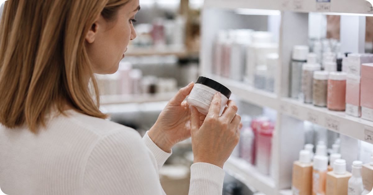 A young woman carefully examines the label on a jar of skincare product while standing in front of shelves stocked with beauty and cosmetic items in a store, highlighting the importance of checking ingredients before purchase.