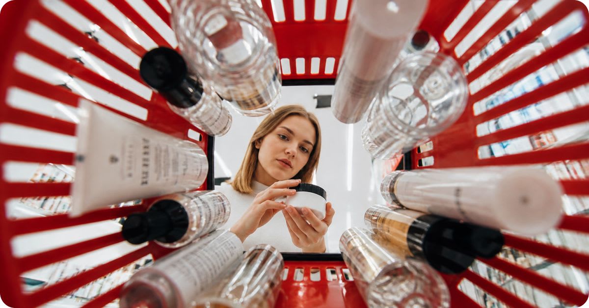 Overhead view from inside a red shopping basket filled with various skincare bottles, tubes, and jars, with a woman looking down into the basket as she inspects her cosmetic products for potentially harmful ingredients.