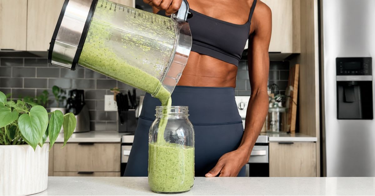Fit woman in athletic wear pouring a fresh green vegetable smoothie from a blender into a glass jar in a modern kitchen — representing the inside-out approach to healthy, glowing skin