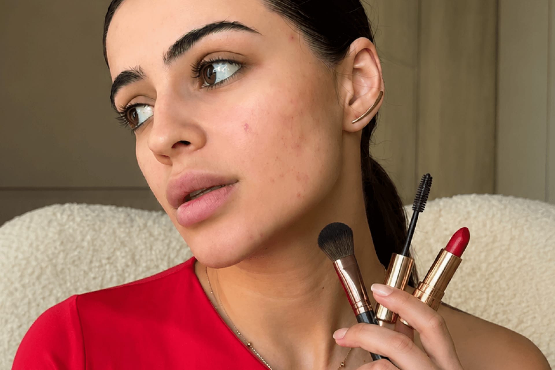 Close-up of a young woman with visible acne and breakouts on her cheek, holding a makeup brush, mascara wand, and red lipstick up to her face, wearing a red top against a soft beige boucle background.