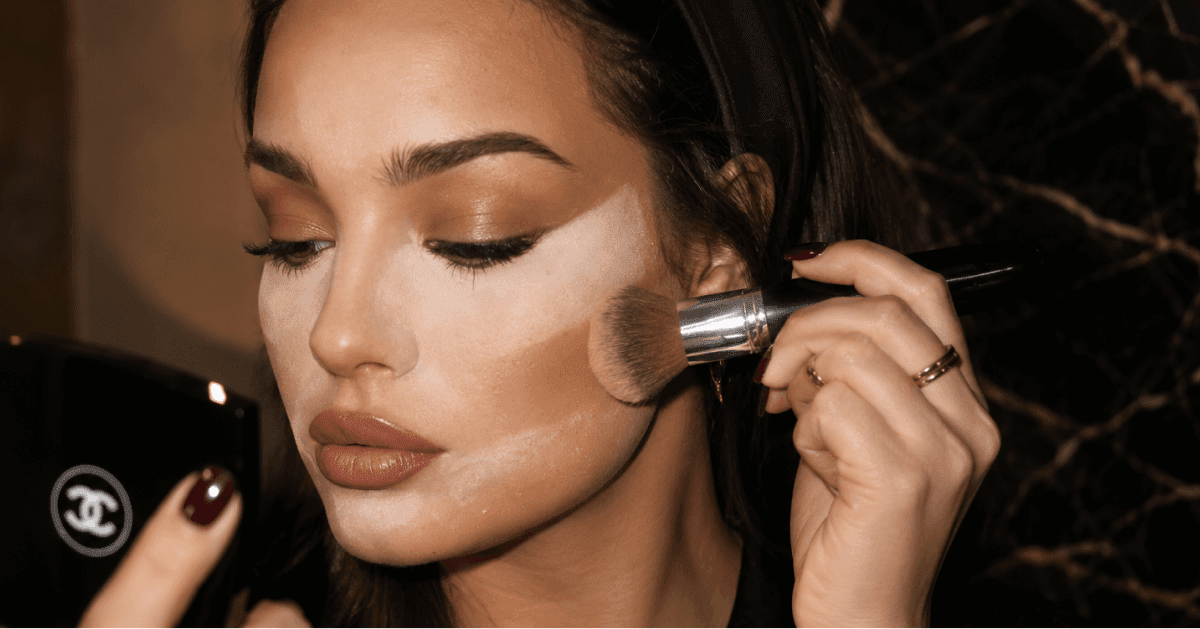 Dark-haired woman sweeping a large fluffy brush across her cheek to apply powder, with a black Chanel compact nearby.