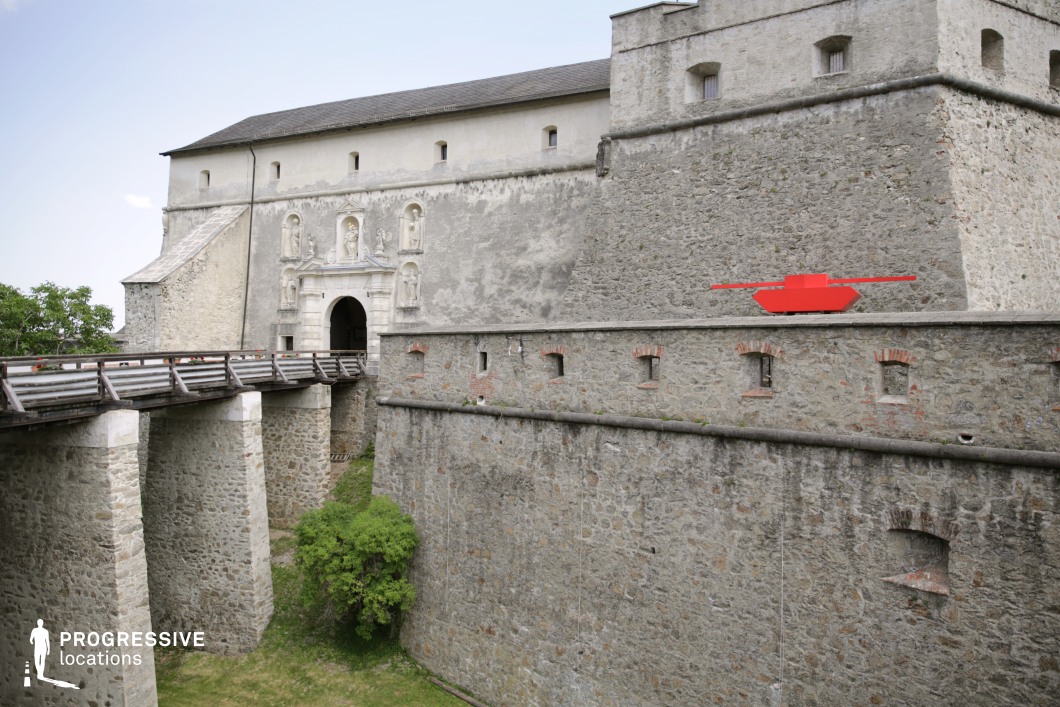 This imposing fortress features thick stone walls with a fortified design, including narrow defensive openings and a high parapet, showcasing a blend of medieval and early modern military architecture. The grand entrance is framed by an ornate sculpted portal, adding a touch of refinement to the otherwise rugged structure. A wooden and stone bridge spans a dry moat, enhancing the location’s fortified appearance. The textured stone surfaces, aged patina, and fortified layout create a historically rich atmosphere. This location is ideal for period dramas, historical battles, or fantasy epics, offering an authentic setting for medieval or war-themed storytelling.