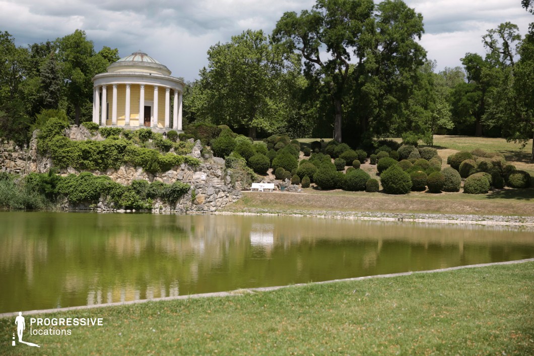 This idyllic landscape features a neoclassical pavilion with a domed roof and white columns, elegantly perched atop a rocky, vine-covered hillside. The structure is framed by lush greenery and meticulously shaped shrubs, contributing to the refined and serene atmosphere. Below, a still lake reflects the surrounding elements, enhancing the symmetry and tranquility of the setting. The combination of classical architecture and natural beauty makes this location ideal for historical dramas, romantic period pieces, or introspective scenes, offering a timeless and elegant backdrop for cinematic storytelling.