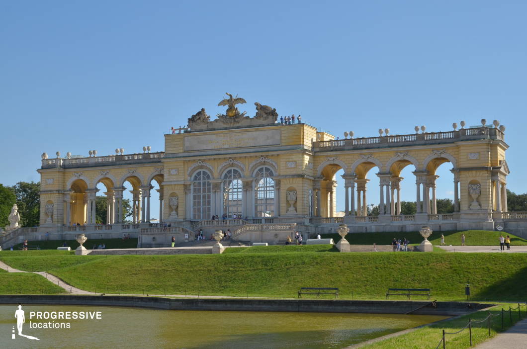 This grand neoclassical pavilion stands atop a gently sloping hill, featuring a symmetrical series of arches supported by ornate columns, intricate stone carvings, and decorative balustrades. The golden-hued facade exudes elegance, crowned by a sculptural crest depicting an imperial eagle, reinforcing its regal character. The foreground showcases a manicured lawn and a reflective pond, enhancing the structure’s stately presence. The open colonnades and raised terrace create a sense of openness and grandeur, making this an ideal location for historical dramas, royal-themed productions, or elegant outdoor sequences. The balanced composition of nature and architecture offers a cinematic backdrop for refined storytelling.