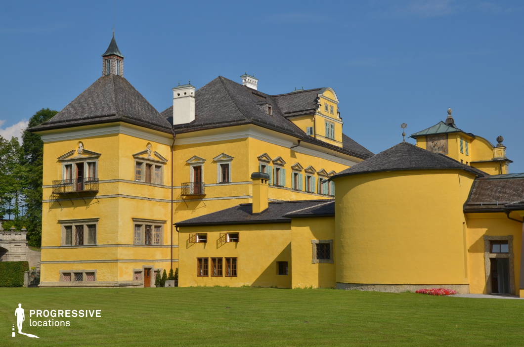 This striking historic estate features a bold yellow facade with elegant architectural details, including arched windows, decorative reliefs, and small wrought-iron balconies. The structure’s distinctive sloped roofs, multiple chimneys, and rounded annexes create an intriguing mix of symmetry and variation. The surrounding manicured lawn enhances the estate’s grandeur, offering an open, scenic setting. The combination of bright colors and traditional European design makes this location ideal for period dramas, aristocratic settings, or whimsical fantasy films. Its regal yet inviting character provides a versatile backdrop for elegant and refined storytelling.