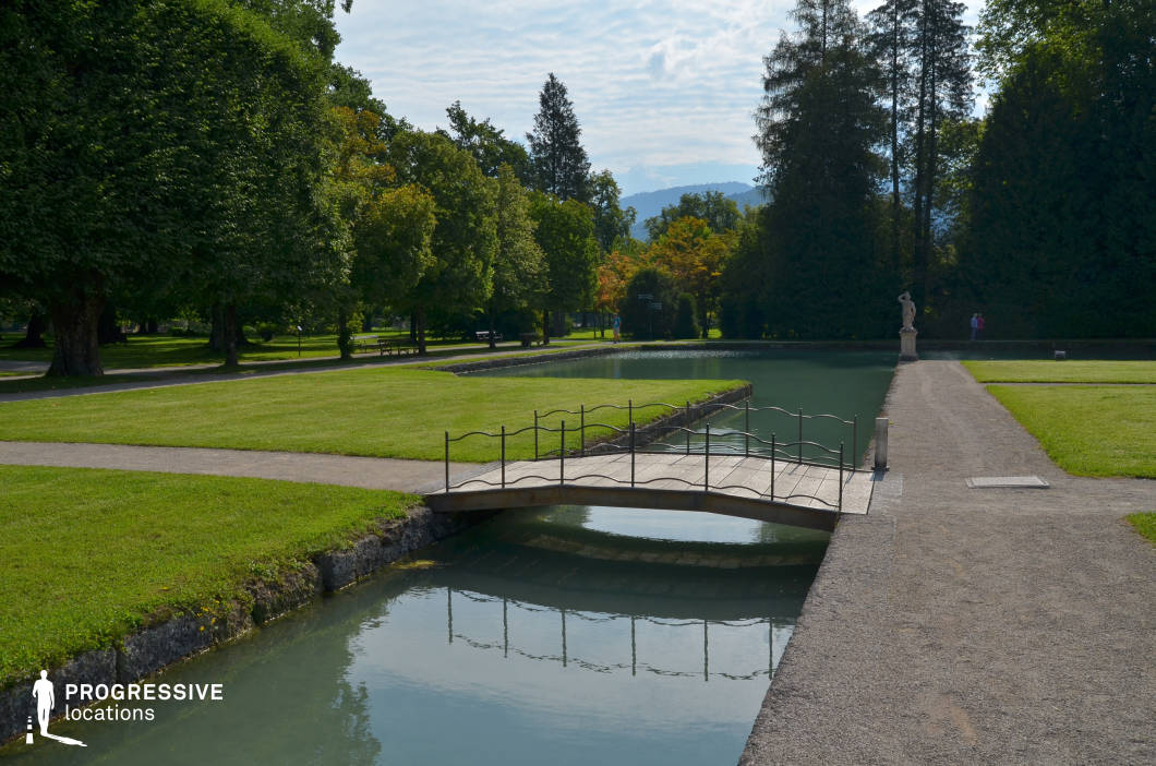 This picturesque garden setting features a small arched wooden bridge with delicate wrought-iron railings spanning a narrow canal. The water reflects the bridge and surrounding greenery, adding depth and symmetry to the scene. The manicured lawns, neatly arranged gravel paths, and a distant classical statue contribute to a refined and serene atmosphere. Towering trees provide natural framing, while the distant mountains in the background enhance the scenic composition. This location is ideal for filming period dramas, romantic scenes, or introspective moments in a tranquil, aristocratic garden, offering a visually rich and timeless setting.