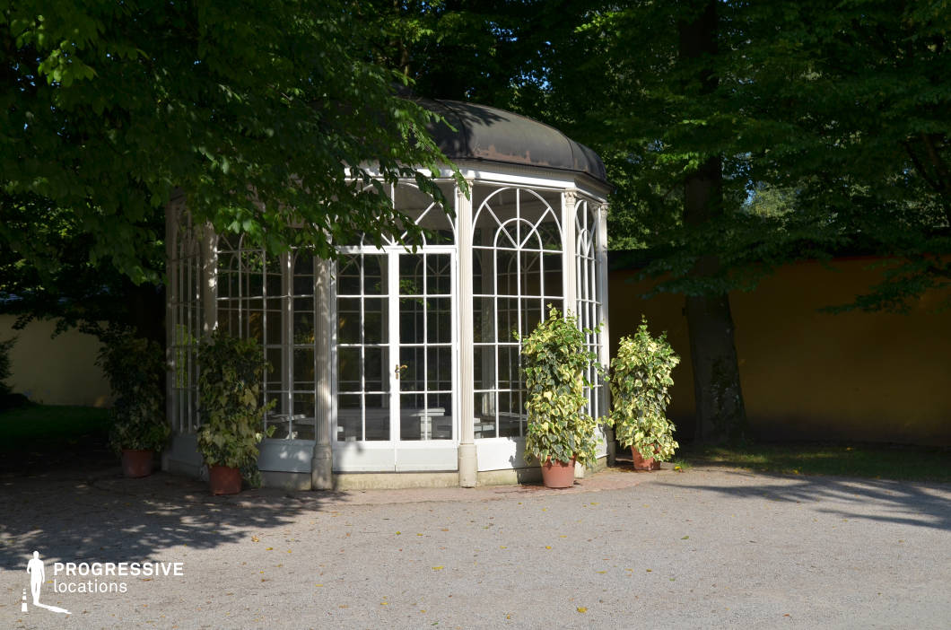 This delicate, octagonal glass gazebo features intricate arched window frames and a domed roof, exuding a timeless charm. Positioned within a lush, tree-lined garden, it is partially shaded by overhanging branches, creating a dappled light effect on the ground. The soft, neutral tones of the structure contrast with the surrounding greenery, enhancing its romantic and classical aesthetic. The setting is ideal for filming period dramas, romantic encounters, or contemplative scenes, providing a refined and intimate atmosphere. The transparent walls offer dynamic framing opportunities, making the gazebo a visually striking element for both exterior and interior shots.