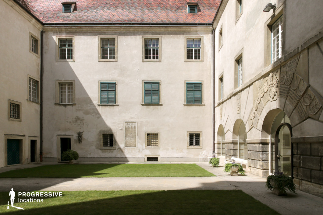 This enclosed courtyard features aged stone facades, arched entryways, and a symmetrical arrangement of windows with wooden shutters, evoking a historical European ambiance. The weathered plaster walls and intricately carved reliefs above the arches add a sense of antiquity, while the red-tiled roof complements the muted tones of the structure. A well-maintained lawn and minimal decorative elements contribute to the serene, enclosed atmosphere. The location’s soft interplay of light and shadow enhances its depth and period aesthetic. This setting is ideal for historical dramas, period thrillers, and medieval or Renaissance-themed films, offering an authentic backdrop for aristocratic, monastic, or secretive intrigue scenes.