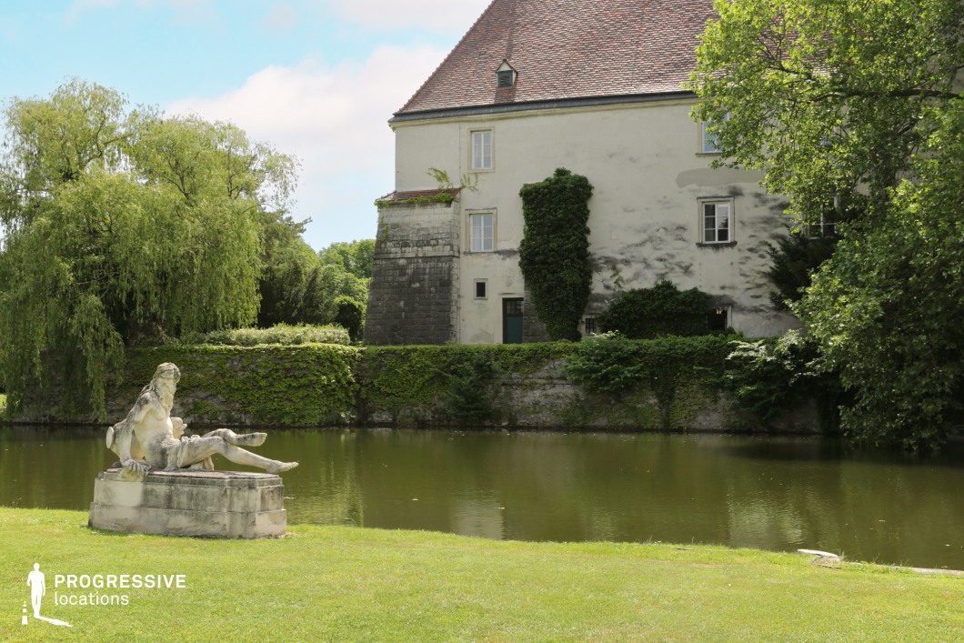 This serene setting features a historic building with a steep red-tiled roof and aged plaster walls, partially covered in creeping ivy, exuding a timeless elegance. A stone retaining wall, similarly adorned with vegetation, encircles a reflective body of water, enhancing the secluded and classical atmosphere. A weathered sculpture on a stone pedestal adds an artistic focal point, reinforcing the historical and cultural richness of the location. The lush greenery and carefully manicured lawn contrast beautifully with the rustic textures of the architecture. This location is ideal for period dramas, romantic storytelling, and fantasy films, offering a picturesque backdrop for aristocratic gatherings, poetic solitude, or mystical narratives.