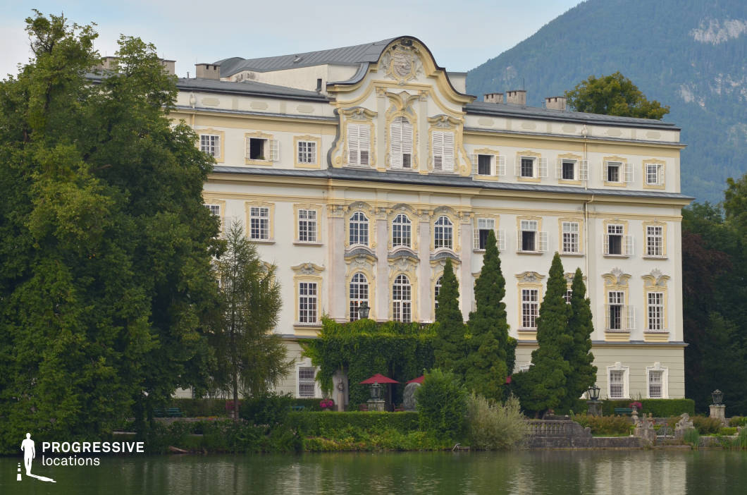 This grand Baroque-style mansion is set against a serene lake, framed by lush greenery and rolling hills in the background. The pale cream and gold façade is adorned with intricate stucco details, arched windows, and decorative shutters, exuding an air of aristocratic refinement. The lower terrace, partially covered in ivy, features manicured gardens, ornamental statues, and shaded seating areas, enhancing the sense of elegance and tranquility. The reflective water surface in the foreground adds depth and symmetry to the scene. This location is ideal for filming historical dramas, period romances, or mystery thrillers, offering a sophisticated and secluded setting with timeless charm.