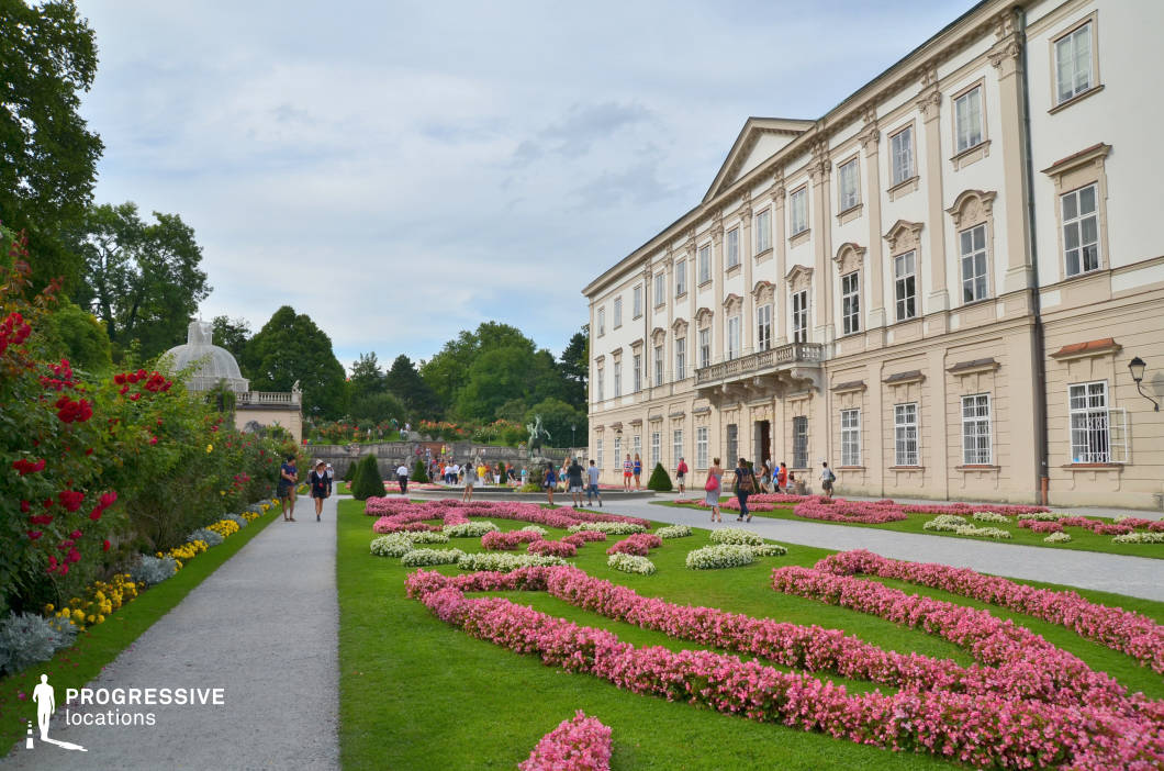 This grand palace features a symmetrical Baroque-style facade with intricate detailing, large windows, and a stately entrance. In the foreground, a meticulously designed formal garden showcases swirling flower arrangements in vibrant colors, bordered by manicured hedges and gravel pathways. A classical dome and stone balustrades in the background add depth and historical character to the setting. The balance of architectural elegance and lush greenery creates a refined and sophisticated atmosphere. This location is ideal for period dramas, romantic films, and historical adaptations, offering a regal and timeless backdrop for aristocratic settings, formal gatherings, or idyllic outdoor sequences.