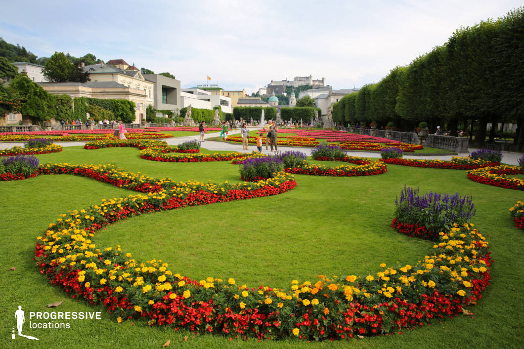 This meticulously designed Baroque garden features vibrant, swirling flower beds arranged in intricate patterns across a lush green lawn. The symmetrical layout is complemented by a tree-lined promenade and classical sculptures, enhancing the elegance of the setting. In the background, a mix of historic and modern architecture is visible, with a grand fortress perched atop a hill, adding a dramatic focal point. The blend of rich colors, precise landscaping, and historic grandeur makes this an ideal filming location for period dramas, romance films, and refined, elegant commercial productions, offering a timeless and picturesque European atmosphere.