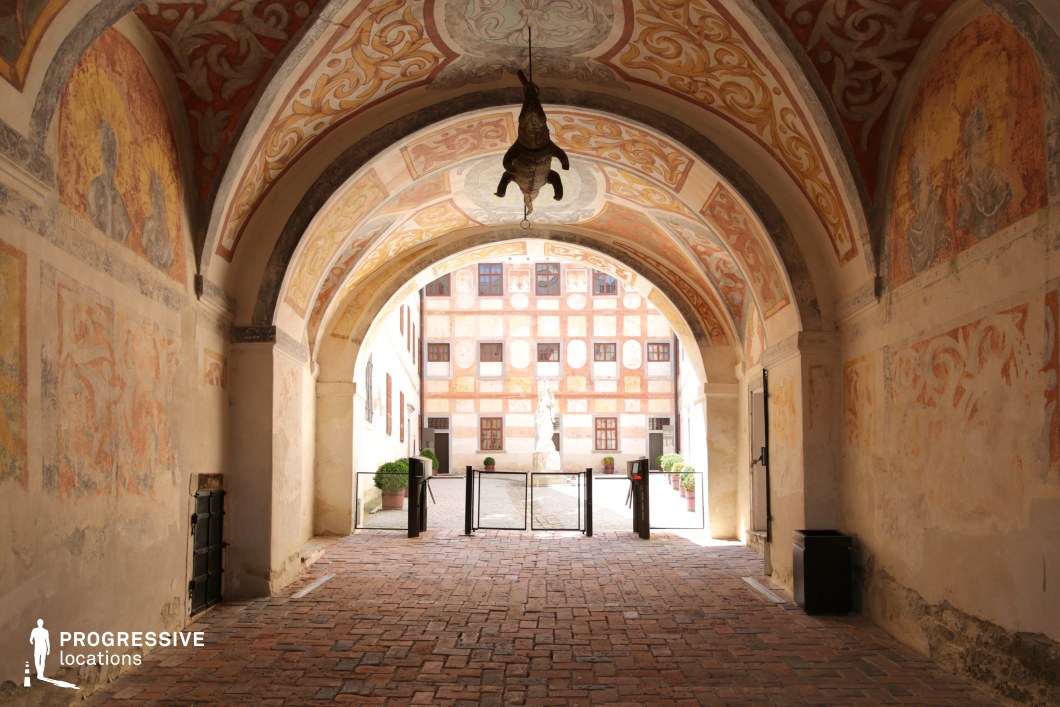 This atmospheric arched passageway, adorned with faded frescoes and intricate ornamental patterns, frames a sunlit courtyard beyond. The aged plaster walls and warm-toned brick flooring enhance the historic character of the space, while the vaulted ceiling, decorated with elegant motifs, adds depth and grandeur. A striking focal point is the unusual suspended taxidermy, adding an element of intrigue. The courtyard visible in the background features a symmetrical façade with painted geometric patterns, a central statue, and potted greenery, reinforcing the sense of classical European heritage. This location is ideal for filming historical dramas, mystery thrillers, or medieval-inspired narratives, offering a visually rich and immersive old-world setting.