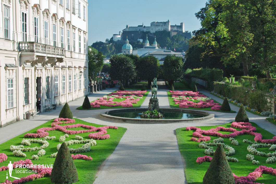 This exquisite baroque garden presents a meticulously landscaped setting with symmetrically arranged flower beds, sculpted hedges, and a central fountain, all framed by a stately classical building with ornate facades and tall windows. The manicured pathways lead the eye toward a breathtaking historic fortress atop a hill in the background, adding a sense of grandeur and timelessness. The combination of refined architecture, lush greenery, and distant medieval structures creates a visually striking contrast, making this location ideal for historical dramas, romantic period pieces, or regal-themed productions. Its balanced composition and vibrant colors offer a sophisticated and cinematic backdrop.