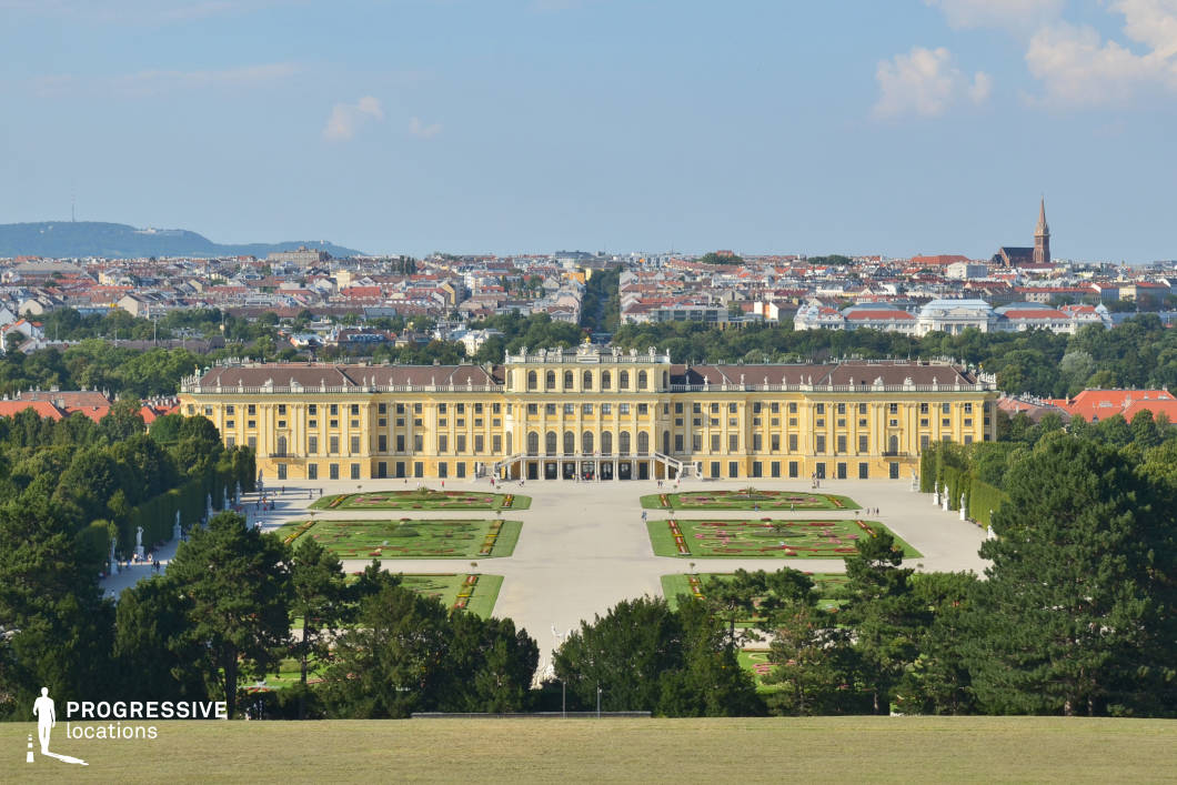 This grand palace stands as a striking example of baroque architecture, with its symmetrical facade, ornate roofline, and extensive gardens that extend outward in precise geometric patterns. The golden-hued exterior contrasts elegantly with the surrounding greenery, while the wide central courtyard emphasizes the building’s monumental scale. The background features a dense cityscape with red-roofed buildings and distant hills, adding depth and historical context. This location is ideal for filming period dramas, royal narratives, or historical epics, providing a regal and cinematic atmosphere. The elevated vantage point offers sweeping panoramic shots, making it perfect for establishing sequences or majestic arrival scenes.