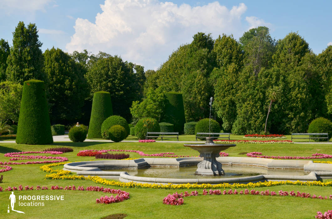 This meticulously designed garden features a central stone fountain surrounded by precisely arranged flower beds in vivid patterns. The lush green lawns are adorned with sculpted topiary trees, creating a sense of order and refinement. In the background, dense trees provide a natural border, adding depth and contrast to the structured landscape. The symmetrical layout and classical elements evoke a regal and serene atmosphere, making this location ideal for filming historical dramas, romance films, and period pieces. The harmonious blend of greenery, water features, and ornamental design provides a visually rich backdrop for elegant outdoor scenes or leisurely promenade sequences.