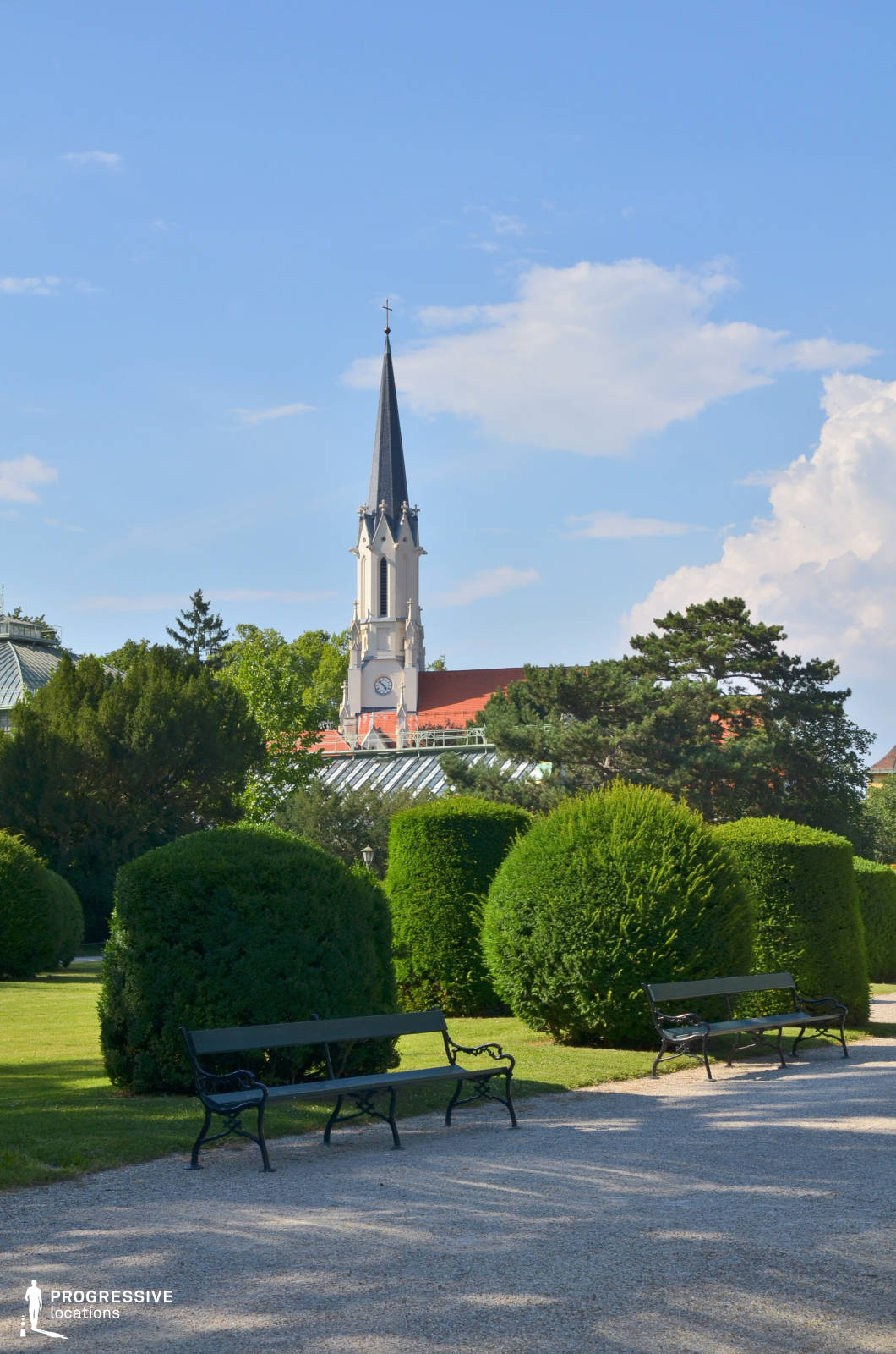 This picturesque setting features a meticulously maintained garden with neatly trimmed hedges and a gravel pathway lined with classic wrought-iron benches. The composition is framed by a striking Gothic-style church with a pointed spire, delicate architectural detailing, and a clock face, standing against the backdrop of a clear sky. The combination of lush greenery and historic architecture creates a serene and refined atmosphere. This location is ideal for period dramas, romantic narratives, or introspective scenes, offering a visually rich and timeless European aesthetic.