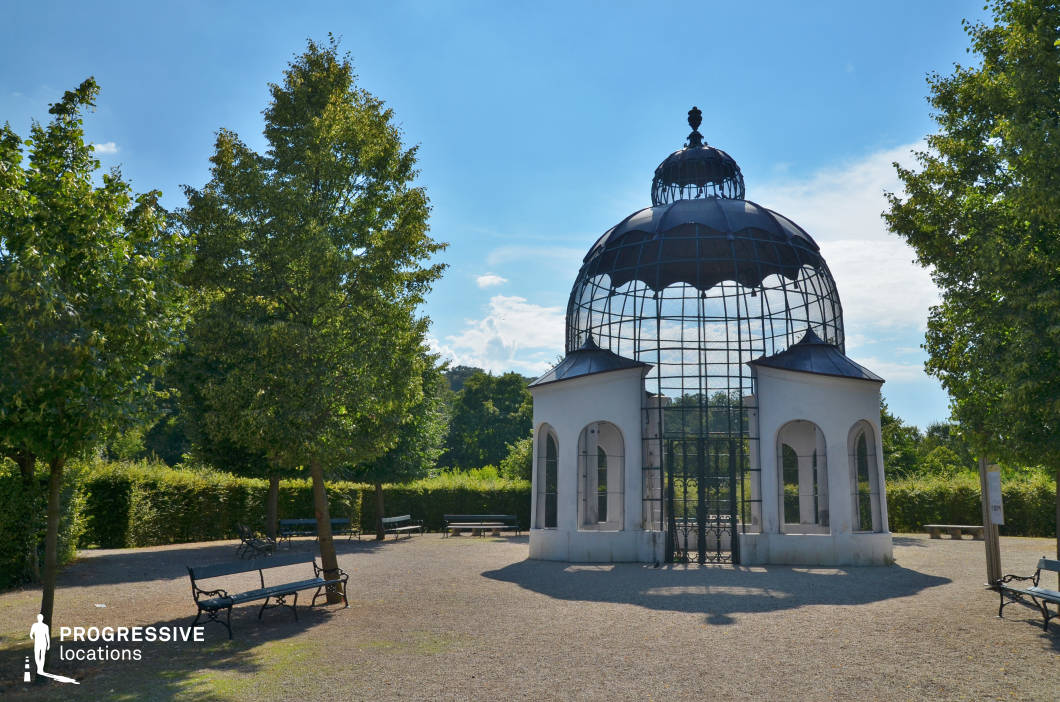 This elegant garden structure features a distinctive domed metal framework atop a classical white pavilion, blending ornamental and functional design. The wrought-iron lattice dome, crowned with a decorative finial, contrasts with the smooth, arched entryways of the supporting pavilion, creating a visually striking focal point. The surrounding garden is meticulously arranged with neatly trimmed hedges, evenly spaced trees, and gravel pathways, contributing to a refined and serene atmosphere. Classic wrought-iron benches add a historical charm to the space. This location is ideal for filming period dramas, romantic scenes, or fantasy narratives, offering a sophisticated and timeless setting with strong European aristocratic influences.