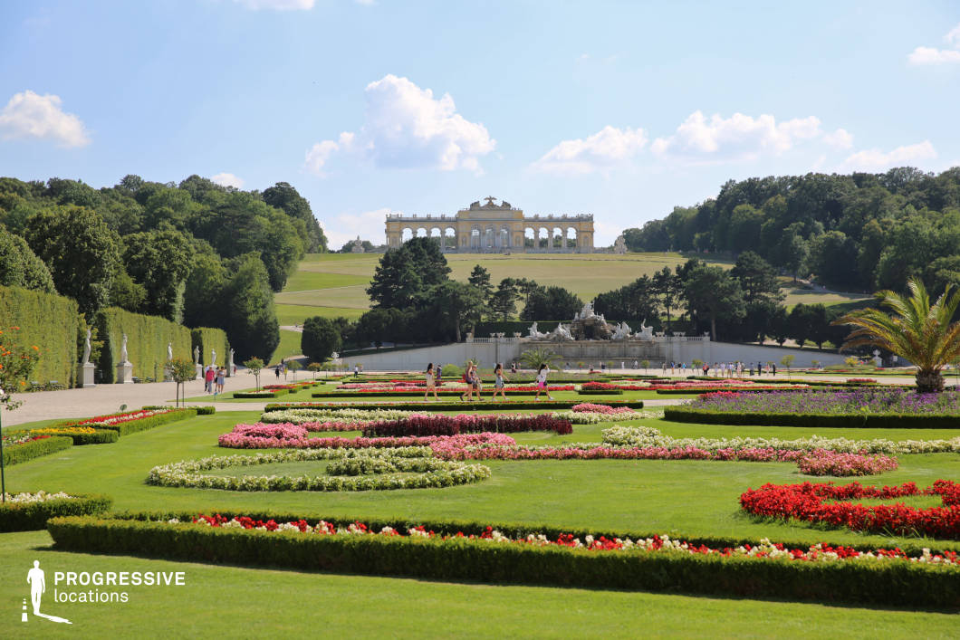 This grand palace garden showcases meticulously designed floral arrangements, symmetrical hedges, and an expansive green lawn leading towards a classical hilltop structure. The structured flower beds, featuring vibrant colors, contrast elegantly with the trimmed pathways and statues lining the perimeter. A sculptural fountain serves as a focal point, adding an element of grandeur to the scene. The gently sloping terrain and distant pavilion create depth, making this an ideal location for period dramas, royal settings, or romantic cinematic sequences requiring a refined and aristocratic ambiance.