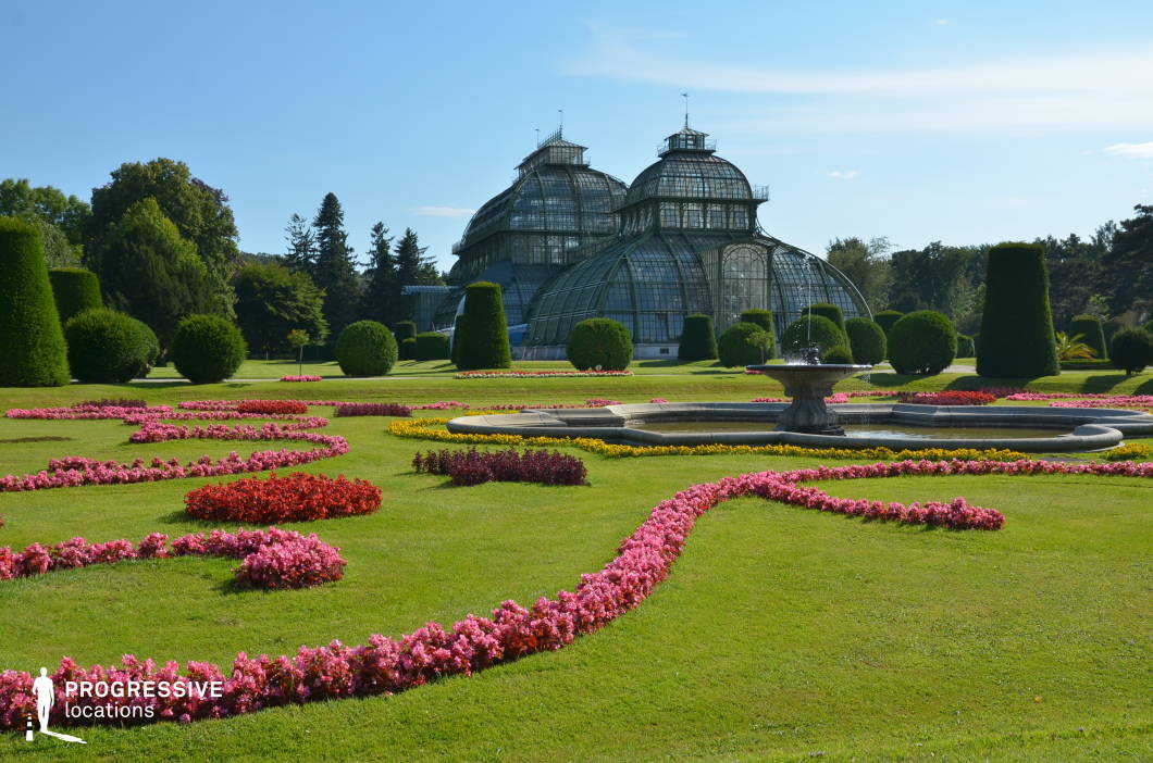 This elegant botanical garden features a meticulously designed landscape with intricate floral arrangements, precisely trimmed topiaries, and a grand ornamental fountain. At the center stands a magnificent historic glasshouse with intricate ironwork and arched domes, embodying classic greenhouse architecture. The vast, well-manicured lawn enhances the symmetry and visual harmony of the setting. The combination of structured greenery and decorative floral patterns creates a refined yet serene atmosphere. This location is ideal for period dramas, romantic films, or historical documentaries, offering a lush and sophisticated backdrop for elegant gatherings, introspective moments, or scenes evoking classical European luxury.