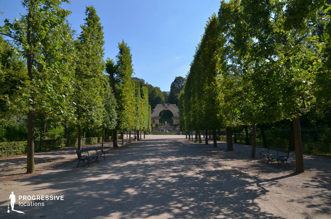 This stately promenade is framed by symmetrically arranged, meticulously pruned trees, creating a natural corridor leading toward an ornate classical ruin. The wide gravel pathway, lined with elegant wrought-iron benches, enhances the formal garden atmosphere, evoking a sense of timeless sophistication. The distant architectural focal point, a grand stone arch with intricate details, adds depth and grandeur to the scene. The harmonious balance of nature and classical design makes this location ideal for period dramas, historical epics, and contemplative sequences, offering a refined and cinematic setting for promenades, secretive meetings, or introspective character moments.