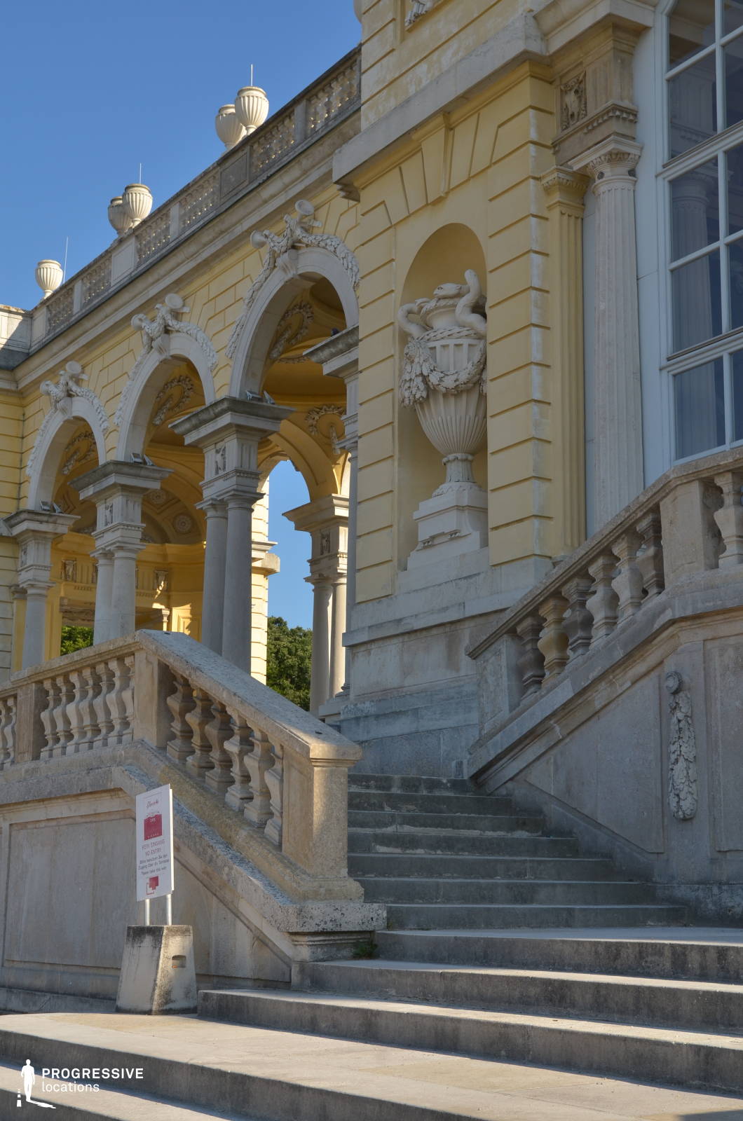 This grand Baroque terrace features elegant arched colonnades adorned with intricate sculptural details and classical motifs. The pale yellow façade is accented by white stone reliefs, including decorative urns and garlands, which enhance the structure’s regal aesthetic. A gracefully curved stone staircase with balustrades leads to the upper level, adding depth and movement to the architectural composition. The sunlight casts sharp shadows on the textured surfaces, highlighting the craftsmanship and historical charm of the structure. This location is ideal for filming period dramas, historical narratives, or refined aristocratic settings, offering an opulent and visually rich backdrop.