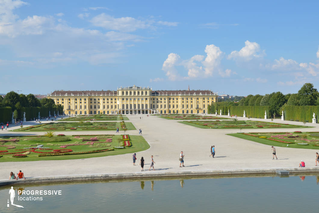 This magnificent palace, with its symmetrical Baroque façade and warm golden tones, serves as the focal point of an expansive formal garden. The meticulously designed landscape features geometric flowerbeds, neatly trimmed hedges, and classical statues lining the wide gravel pathways, all reinforcing the grandeur of the estate. A reflective water feature in the foreground adds depth and balance to the composition. The vast open space, framed by lush greenery, enhances the regal and timeless quality of the scene. This location is ideal for filming historical epics, period dramas, or romantic narratives, offering a luxurious and visually striking setting.