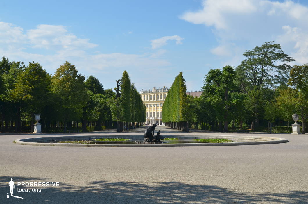 This stately palace avenue is framed by meticulously trimmed trees leading towards the elegant Baroque façade of the grand estate in the distance. A central ornamental fountain with classical sculptures serves as a refined focal point, adding depth and symmetry to the composition. The vast gravel courtyard enhances the sense of openness and formality, while the surrounding dense greenery creates a serene, enclosed atmosphere. This location is ideal for filming period dramas, historical narratives, or aristocratic settings, offering a sophisticated and timeless backdrop for regal and contemplative scenes.