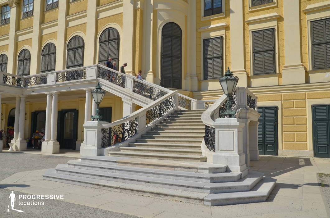 This elegant Baroque-style staircase, framed by ornate wrought-iron railings and flanked by decorative lanterns, leads to the upper level of a historic palace. The building's yellow façade, tall arched windows with dark shutters, and classical columns contribute to an opulent, aristocratic atmosphere. The structure’s symmetry and refined detailing evoke a sense of prestige and grandeur, making it a visually striking backdrop. This location is ideal for filming period dramas, historical narratives, or regal-themed productions, perfectly suited for scenes depicting aristocratic gatherings, royal entrances, or dramatic confrontations in a lavish setting.