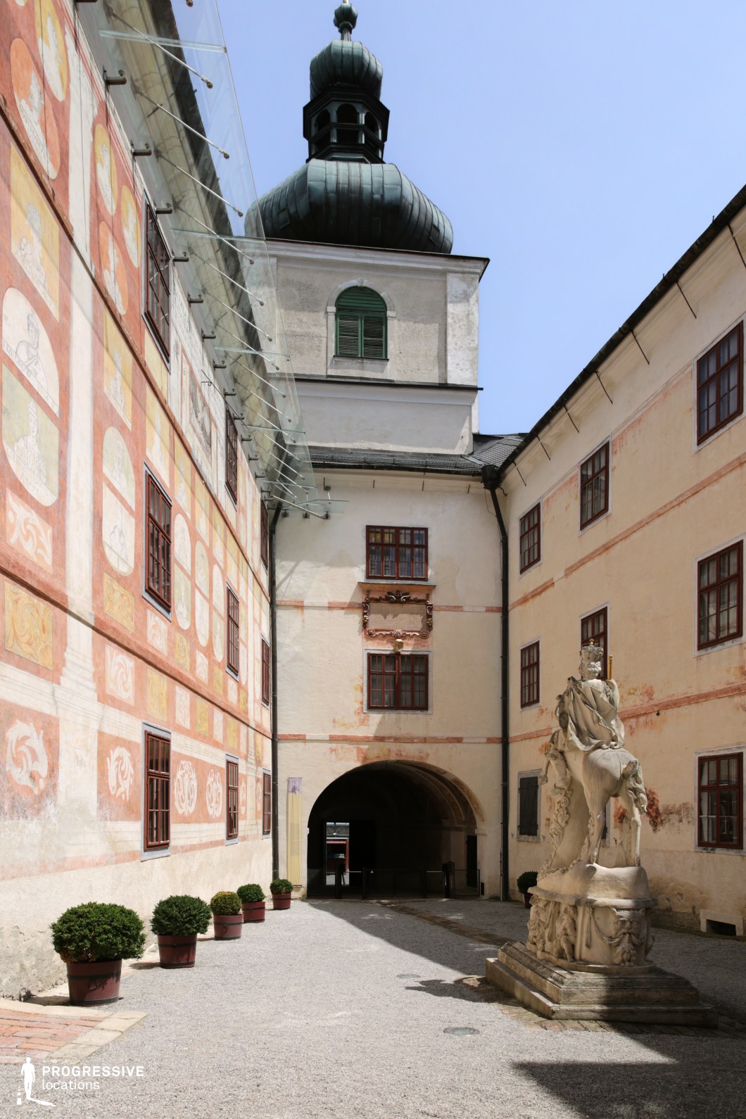 This enclosed courtyard exudes historical charm, featuring aged plaster walls adorned with faded frescoes and a patina-covered tower with a distinctive onion dome. The arched passageway leads into the shaded depths of the structure, enhancing the sense of mystery and antiquity. A weathered stone statue stands prominently, contributing to the classical European aesthetic. The contrasting textures of smooth stone, aged paint, and rough gravel create a visually rich environment. This location is ideal for filming historical dramas, period thrillers, or medieval-themed narratives, offering an authentic and immersive setting with a timeless atmosphere.