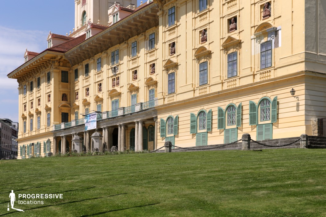 This elegant Baroque palace features a richly decorated yellow façade adorned with intricate architectural details, including sculpted busts, ornamental window pediments, and decorative moldings. The contrasting green shutters and large arched windows add a sense of refinement, while the colonnaded terrace enhances its grandeur. A gently sloping, well-manicured lawn in the foreground provides an open, stately approach to the building. This location is ideal for historical films, period dramas, or aristocratic settings, offering an authentic backdrop for royal gatherings, diplomatic encounters, or sophisticated high-society scenes.