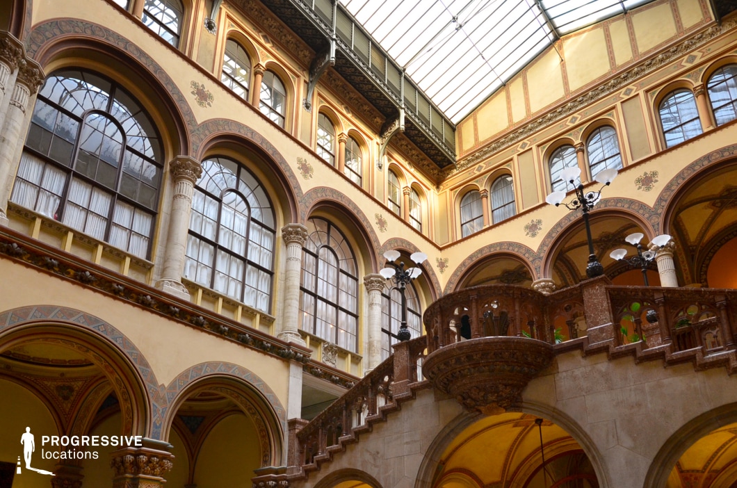 This stunning atrium showcases a harmonious blend of Renaissance and Neo-Gothic architectural elements, featuring soaring arched windows, intricately carved stone balustrades, and elegantly detailed columns. A central staircase with a richly decorated railing adds depth and grandeur, leading to an upper-level gallery encircling the space. The glass ceiling allows natural light to illuminate the warm-toned walls, highlighting the ornamental patterns and intricate moldings. The blend of historic elegance and open, airy design makes this location ideal for filming sophisticated period dramas, aristocratic gatherings, or intellectual salons, providing an opulent and refined setting.