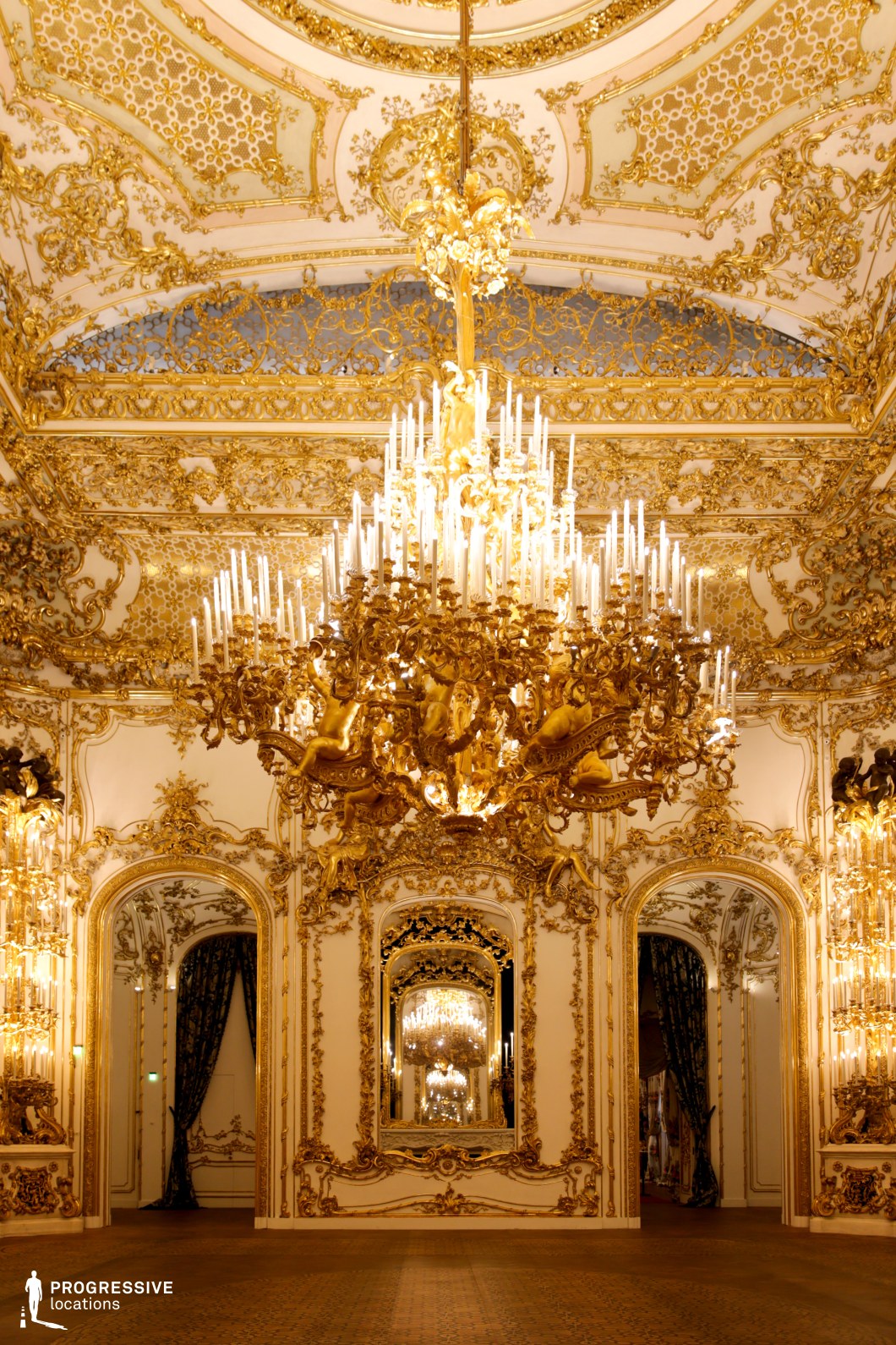 This lavishly decorated Baroque ballroom exudes luxury, featuring intricate gold leaf detailing on the walls and ceiling, elaborate mirrored panels, and ornate archways. The centerpiece is an extravagant gilded chandelier adorned with sculpted cherubs and numerous candle-shaped lights, casting a warm, regal glow. The harmonious blend of white and gold enhances the sense of grandeur, while the symmetrical design and reflective surfaces create depth and elegance. This location is ideal for filming historical dramas, royal banquets, or high-society gatherings, offering a setting that embodies aristocratic opulence and classic European refinement.