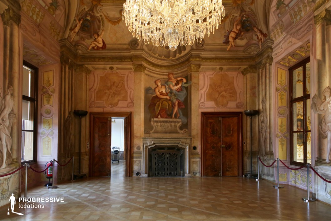 This opulent Rococo-style room is adorned with intricate frescoes depicting classical figures, soft pastel hues, and elaborate gilded details. A grand crystal chandelier hangs from the ceiling, casting a warm glow over the parquet flooring, which features a refined geometric pattern. The richly decorated wooden doors and marble fireplace enhance the historical elegance, while large windows allow natural light to highlight the delicate painted motifs. The ornate yet intimate atmosphere makes this location ideal for filming aristocratic gatherings, period dramas, or scenes of refined intrigue, offering a visually immersive setting for elegant storytelling.