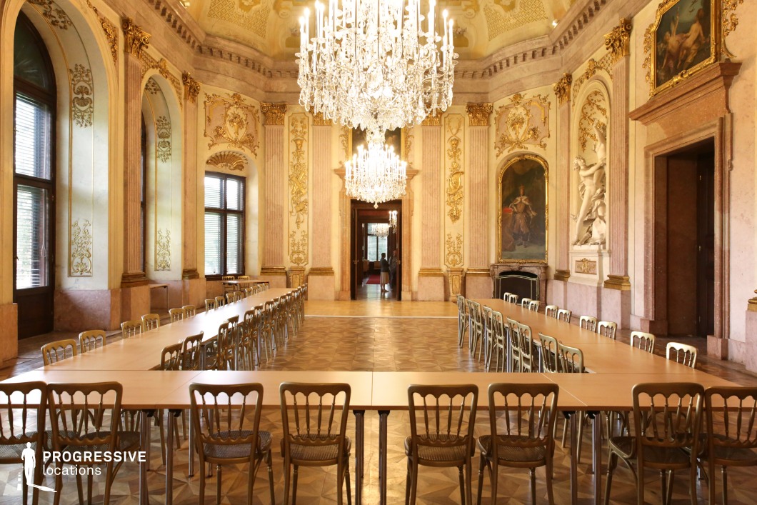 This magnificent hall exudes classical European elegance, featuring high vaulted ceilings adorned with intricate gold detailing, sculpted archways, and large crystal chandeliers that cast a soft, ambient glow. The walls are richly decorated with gilded moldings, painted frescoes, and sculptural reliefs, enhancing the regal atmosphere. The parquet flooring and long banquet-style tables create a structured yet opulent setting, making this space ideal for period dramas, aristocratic gatherings, or high-society political intrigue. The blend of historic grandeur and refined symmetry provides a cinematic backdrop for sophisticated storytelling.