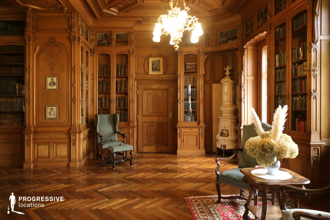 This richly detailed library exudes classical refinement, featuring floor-to-ceiling wooden bookshelves filled with aged volumes. The warm-toned wood paneling, intricate carvings, and coffered ceiling contribute to the space’s distinguished character. A herringbone parquet floor adds to the timeless elegance, while plush green upholstered chairs and a traditional ceramic stove enhance the cozy, intellectual ambiance. The interplay of soft natural light and antique furnishings makes this setting ideal for period dramas, historical thrillers, or refined character-driven narratives, offering an atmosphere of knowledge, sophistication, and intrigue.