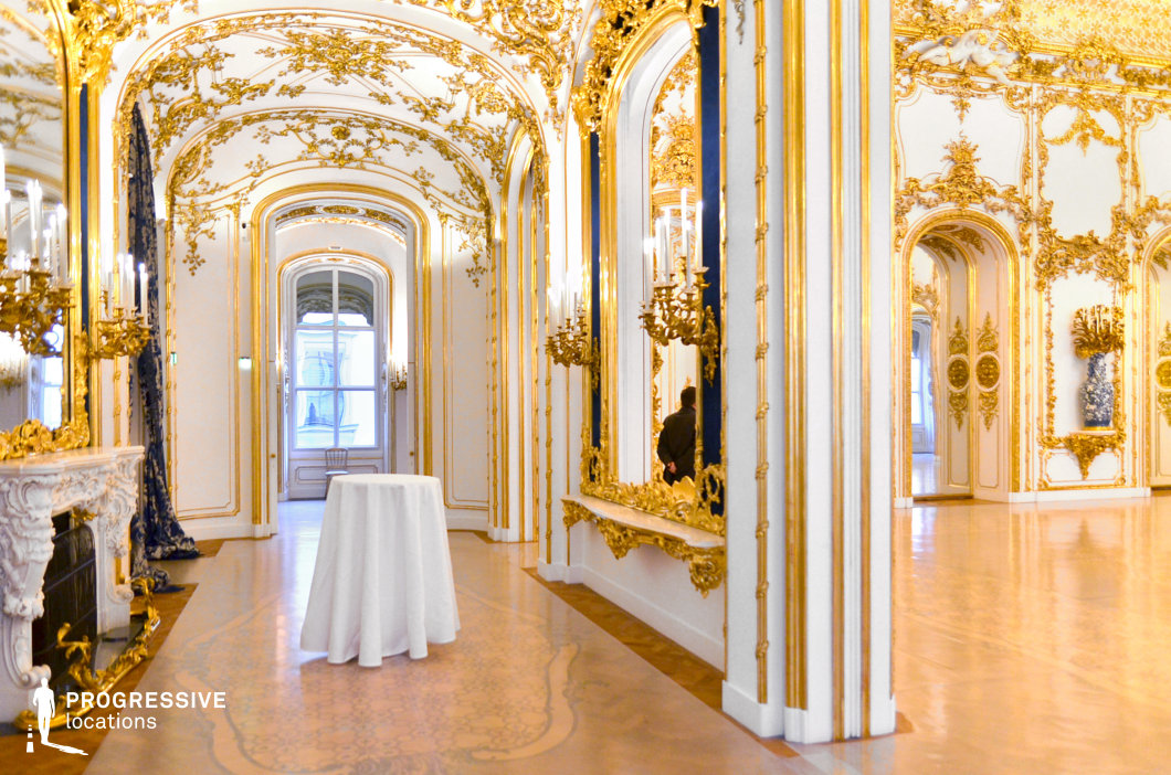 This opulent Rococo-style hallway exudes luxury, with its elaborately gilded moldings, intricate ceiling reliefs, and gracefully arched doorways. The walls are adorned with gold-framed mirrors and delicate sconces, reflecting the warm glow of candle-like lighting. The polished parquet flooring features ornamental inlays, enhancing the regal ambiance. A white marble fireplace with elaborate gold detailing adds to the grandeur, while tall arched windows allow natural light to filter through, accentuating the depth and richness of the décor. This location is ideal for filming historical dramas, royal court scenes, or high-society gatherings, offering an authentic and visually immersive setting for period storytelling.