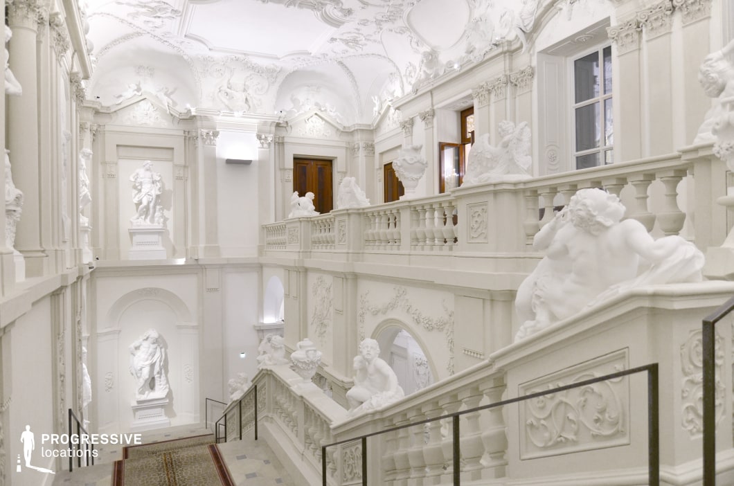 This grand staircase embodies the opulence of Baroque architecture, featuring intricate stucco work, elaborate balustrades, and an abundance of sculptural reliefs. The all-white interior enhances the dramatic interplay of light and shadow, emphasizing the detailed carvings of cherubs, classical figures, and ornamental motifs. The sweeping stairway, framed by high-relief statues set within arched niches, creates a regal and ceremonial atmosphere. The symmetry and depth of the space make it an ideal location for historical dramas, period pieces, and fantasy films, offering a setting befitting royal court scenes, aristocratic gatherings, or mystical encounters in a palatial environment.