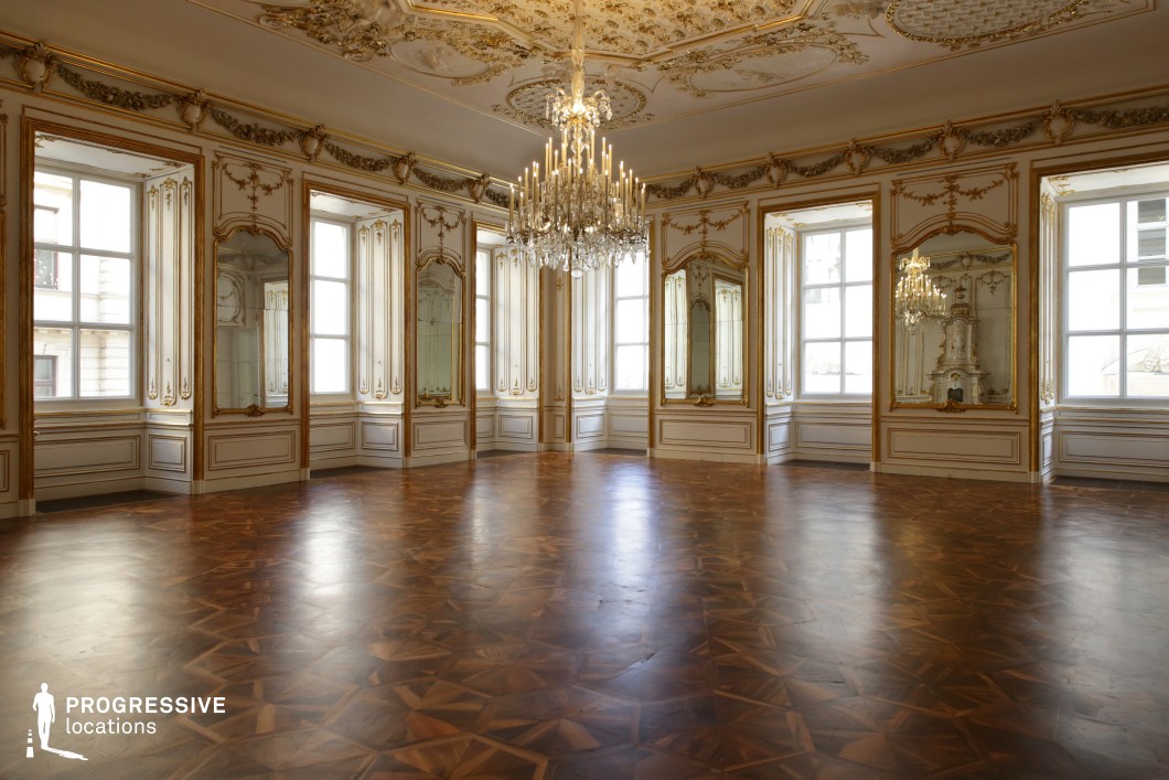 This lavish interior features an exquisite Baroque-style salon adorned with intricate gold moldings, grand mirrors, and an ornately decorated ceiling. A magnificent crystal chandelier hangs from the center, casting reflections across the polished parquet flooring with its rich geometric patterns. Tall windows allow natural light to illuminate the space, enhancing the depth and grandeur of the gold-framed mirrors and delicate plasterwork. The overall aesthetic exudes aristocratic elegance and historical refinement. This location is ideal for filming period dramas, regal ceremonies, or ballroom sequences, offering an authentic and sophisticated setting reminiscent of European palaces.
