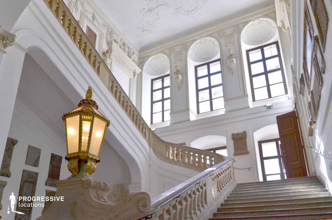 This majestic staircase embodies classical elegance with its sweeping balustrades, intricately carved stonework, and gilded lanterns. The high, arched windows allow natural light to highlight the crisp white stucco reliefs and sculpted busts set within decorative niches. The detailed ceiling moldings and symmetrical architectural elements contribute to the grandeur of the space. The contrast between the warm wooden door and the cool stone surfaces adds depth and refinement. This location is ideal for historical dramas, aristocratic settings, or mystery films, serving as a dramatic backdrop for elegant entrances, secretive meetings, or moments of revelation.