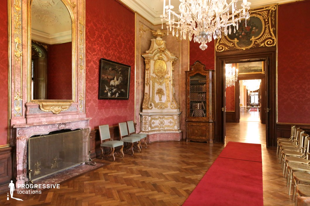 This opulent Rococo-style room exudes aristocratic grandeur, with rich red damask walls, gilded moldings, and a striking marble fireplace adorned with an ornate mirror. The parquet flooring, complemented by a red carpet runner, leads toward an adjoining series of similarly lavish rooms, creating a seamless flow of elegance. Intricately carved wooden furniture, a decorative tiled stove, and a crystal chandelier further enhance the luxurious setting. The space’s warm color palette and exquisite detailing make it an ideal backdrop for historical dramas, period films, and aristocratic intrigue, offering an authentic setting for scenes of power, refinement, or secretive political maneuvering.