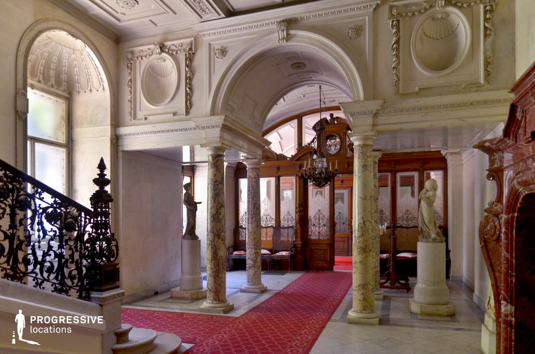 This grand foyer exudes classical refinement with its intricately detailed marble columns, sculpted archways, and decorative wall reliefs. The space is framed by an ornate wrought-iron staircase and a richly carved red marble fireplace, adding to its aristocratic ambiance. A luxurious red carpet guides the eye through the space, leading to an elegant wooden partition with frosted glass panels and intricate etchings, providing a sense of exclusivity. Sculptural elements and soft, natural lighting further enhance the regal atmosphere. This location is ideal for filming period dramas, historical mysteries, or high-society narratives, offering a visually rich and sophisticated setting.