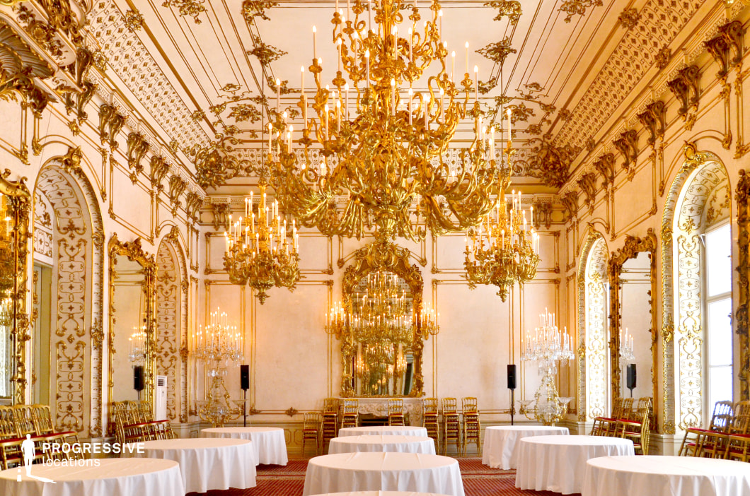 This lavish Baroque ballroom exudes elegance with its intricate gilded moldings, towering mirrors, and ornate chandeliers casting a golden glow throughout the space. The high ceiling is adorned with elaborate detailing, while the arched windows and mirrored walls amplify the room’s grandeur and sense of space. Round tables draped in white linen contribute to the formal and luxurious atmosphere, making it an ideal setting for regal gatherings. The symmetrical arrangement of chandeliers and decorative elements enhances the visual harmony. This location is perfect for filming historical dramas, royal events, or high-society affairs, providing a setting rich in sophistication and old-world charm.